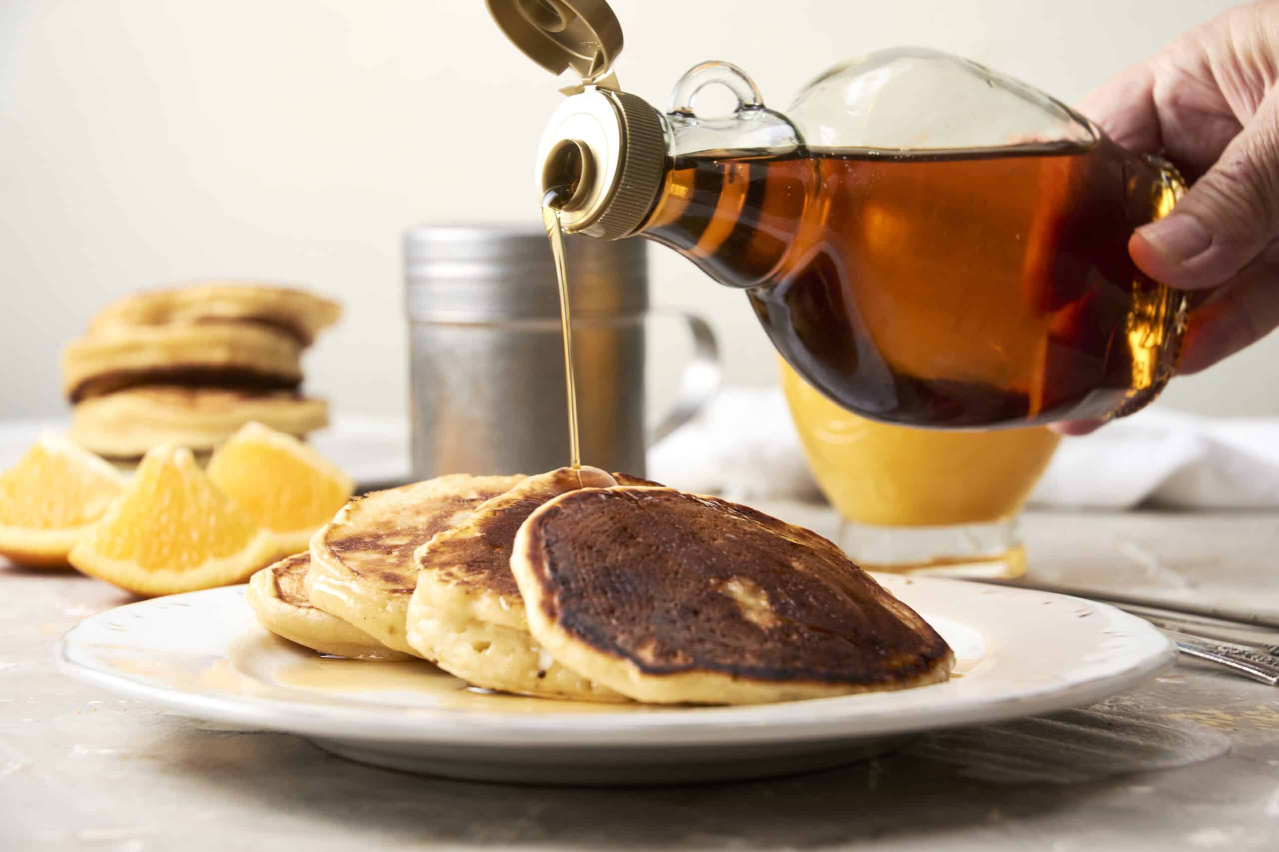 Sweet syrup being poured over a stack of fluffy pancakes with lemon slices in the background, creating a delicious breakfast scene.