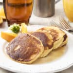 Fluffy pancakes served with lemon wedges, fresh flowers, and breakfast beverages on a white plate. Perfect for a delicious breakfast or brunch, featuring golden-brown pancakes with syrup.