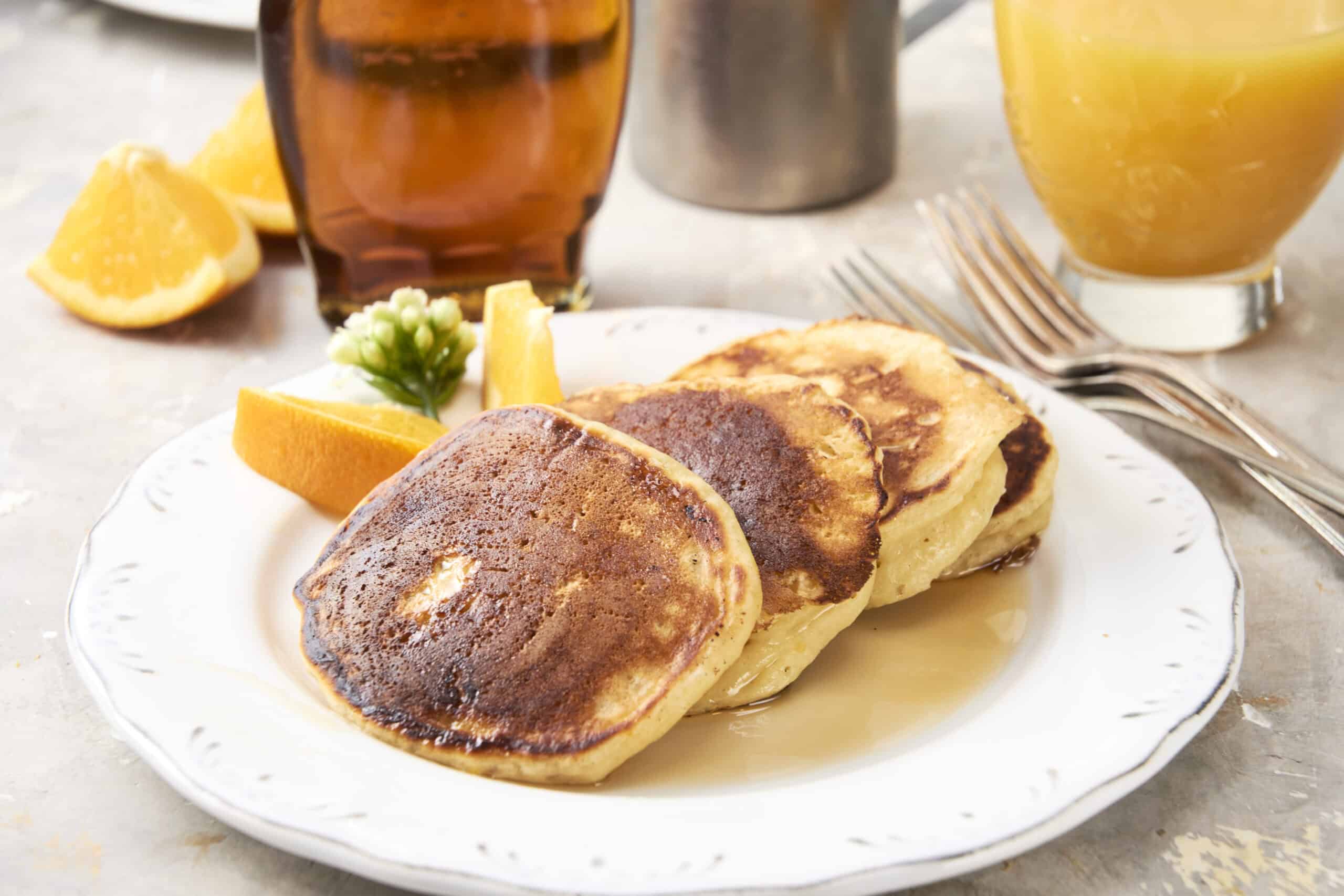 Fluffy pancakes served with lemon wedges, fresh flowers, and breakfast beverages on a white plate. Perfect for a delicious breakfast or brunch, featuring golden-brown pancakes with syrup.