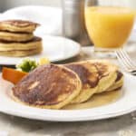 Buttermilk pancakes with crispy edges served with fresh orange slices and syrup on a white plate with a glass of orange juice and a jar of syrup in the background.