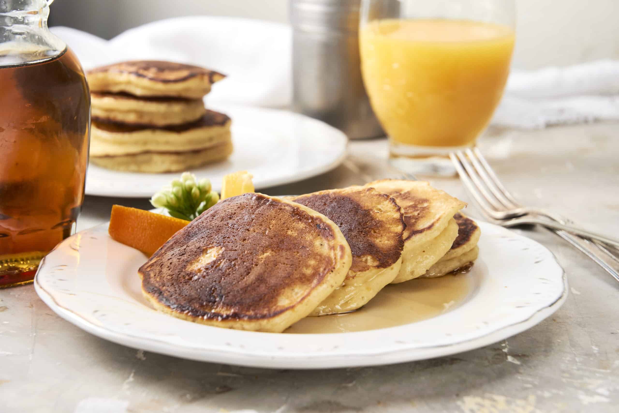 Buttermilk pancakes with crispy edges served with fresh orange slices and syrup on a white plate with a glass of orange juice and a jar of syrup in the background.