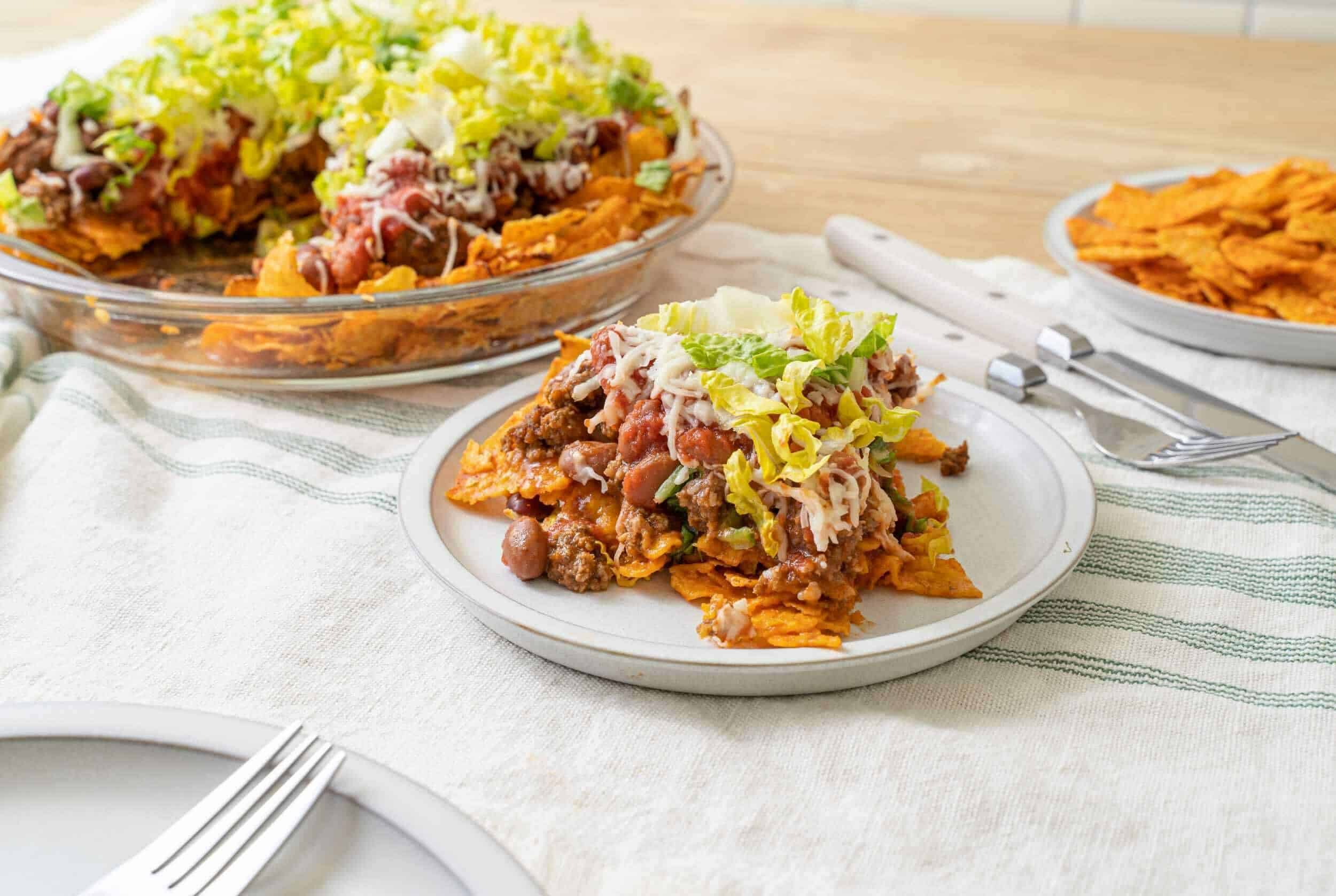 Cheeseburger casserole with shredded cheese, lettuce, ground beef, and tortilla chips on a white plate with a slice served on a white dish.