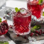 Refreshing pomegranate cocktail with ice cubes and mint in a crystal glass, garnished with mint leaves, surrounded by pomegranate halves, scattered pomegranate seeds, and decorative silver ornaments on a rustic wooden board.