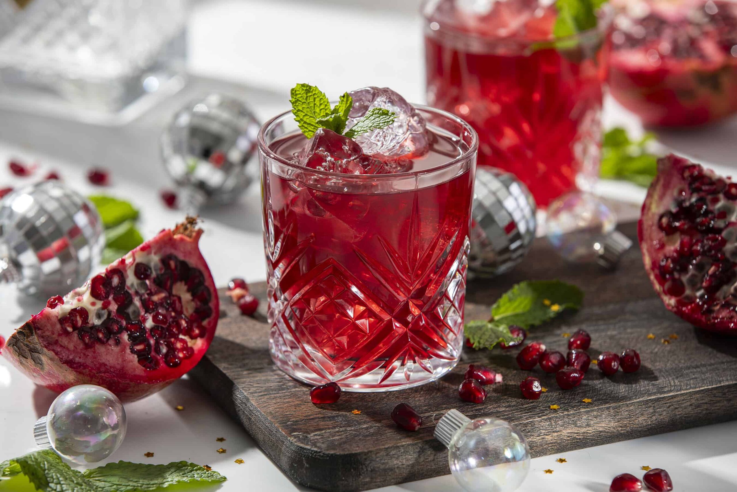 Refreshing pomegranate cocktail with ice cubes and mint in a crystal glass, garnished with mint leaves, surrounded by pomegranate halves, scattered pomegranate seeds, and decorative silver ornaments on a rustic wooden board.