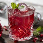 Fresh pomegranate cocktail with ice, mint garnish, and vibrant red color on a rustic wooden surface.