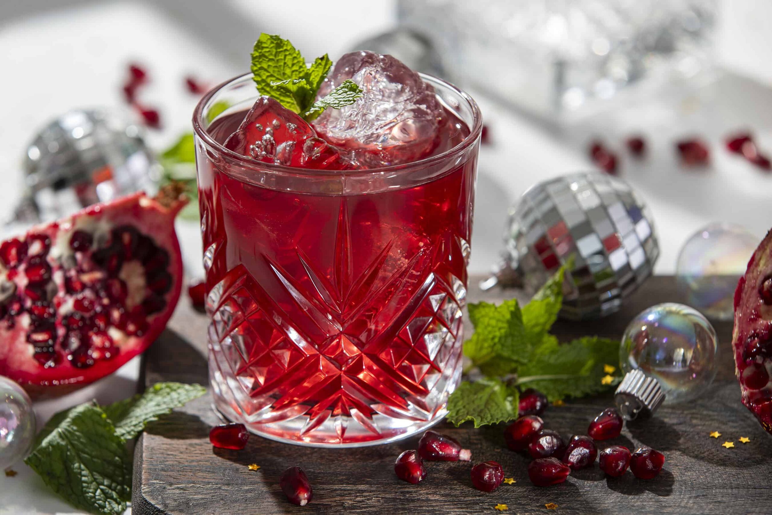 Vivid pomegranate cocktail with ice and garnished with mint leaves, served in a decorative cut glass, surrounded by pomegranate seeds, fresh mint, and holiday ornaments for a festive mood.
