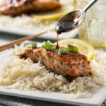 Succulent grilled salmon steak topped with savory sauce, served over fluffy white rice with lemon slices and fresh green onions on a white plate.