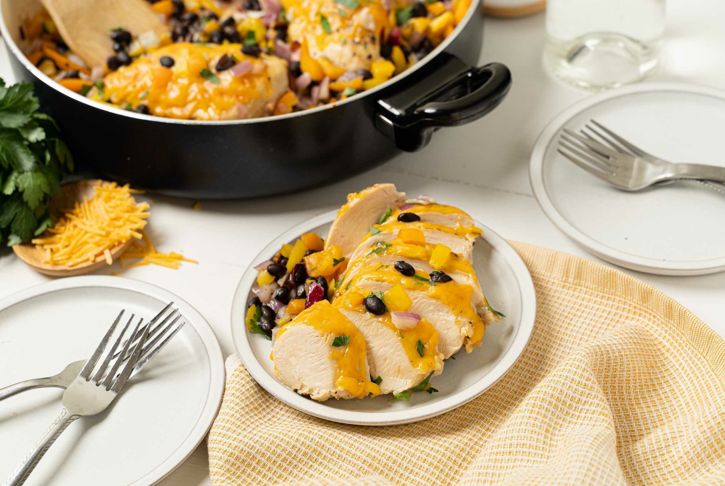 Juicy roasted chicken breast topped with melted cheese, mango salsa, and herbs served on a white plate with a side of black bean salad, perfect for healthy dinner recipes.
