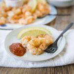 Juicy grilled shrimp with lemon wedge, spicy cocktail sauce, and seasoned black pepper on white plate, served with a fork on a rustic wooden table.