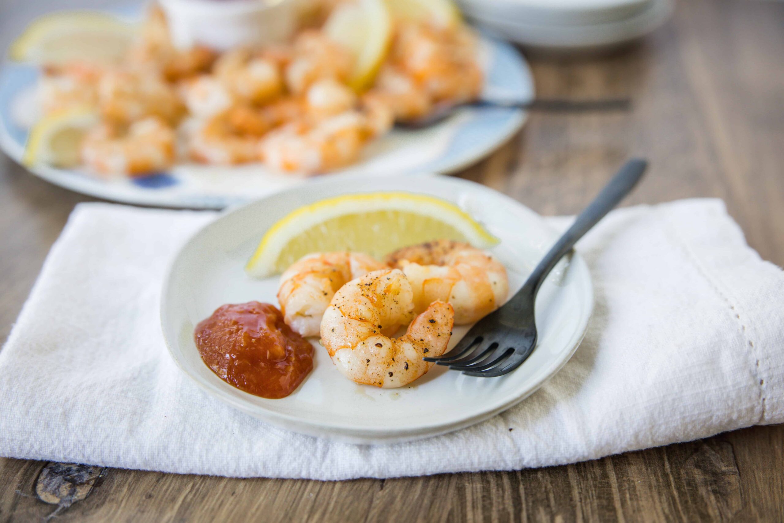 Juicy grilled shrimp with lemon wedge, spicy cocktail sauce, and seasoned black pepper on white plate, served with a fork on a rustic wooden table.