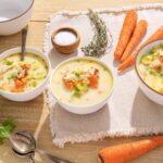 Creamy chicken and vegetable soup with carrots, celery, and peas in white bowls, surrounded by fresh ingredients like whole carrots, celery stalks, thyme, salt, and pepper on a wooden table.