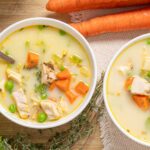 Creamy chicken and vegetable soup with carrots, peas, and herbs in bowls, wooden table, fresh vegetables, and spices for hearty, homemade soup recipe.