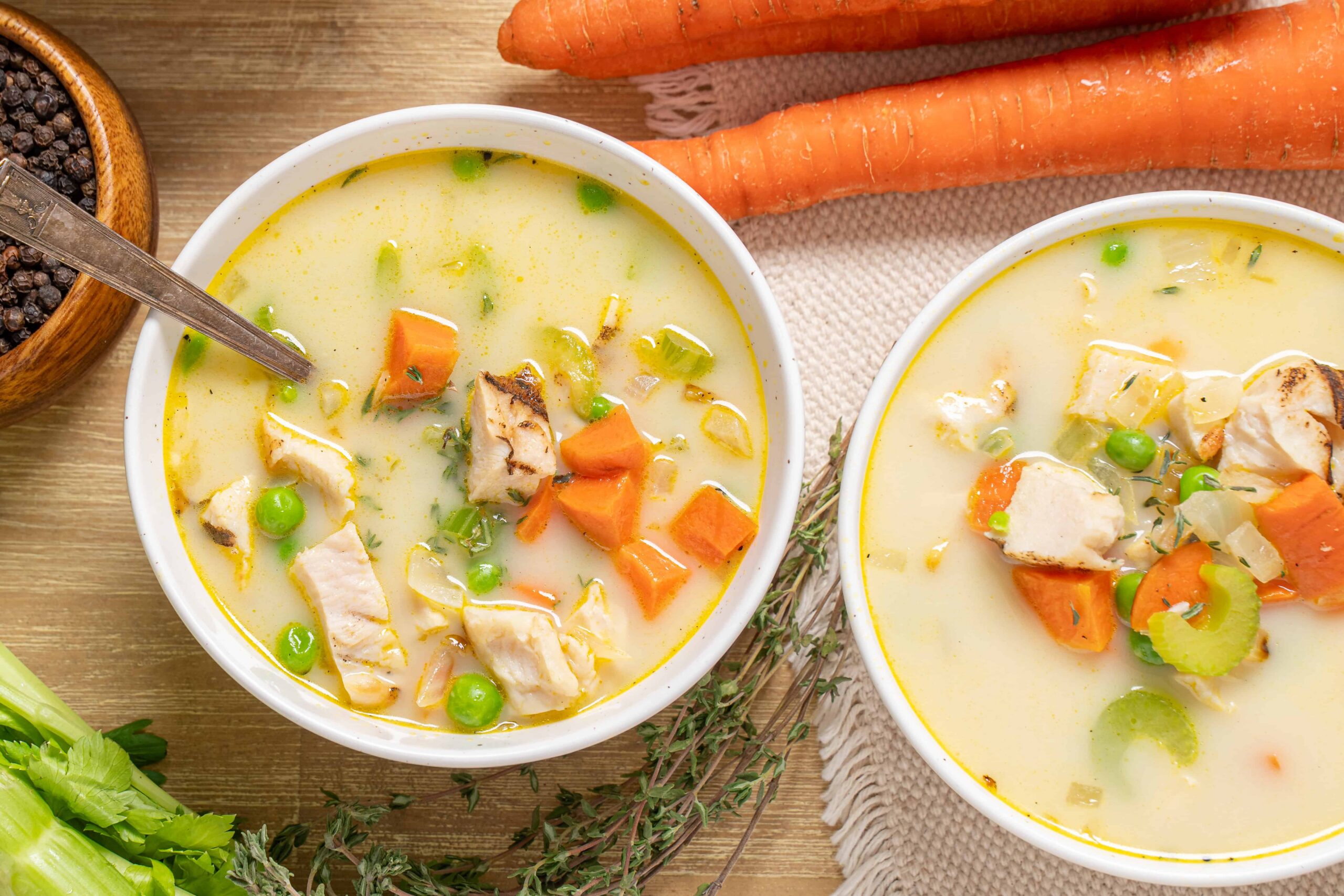 Creamy chicken and vegetable soup with carrots, peas, and herbs in bowls, wooden table, fresh vegetables, and spices for hearty, homemade soup recipe.
