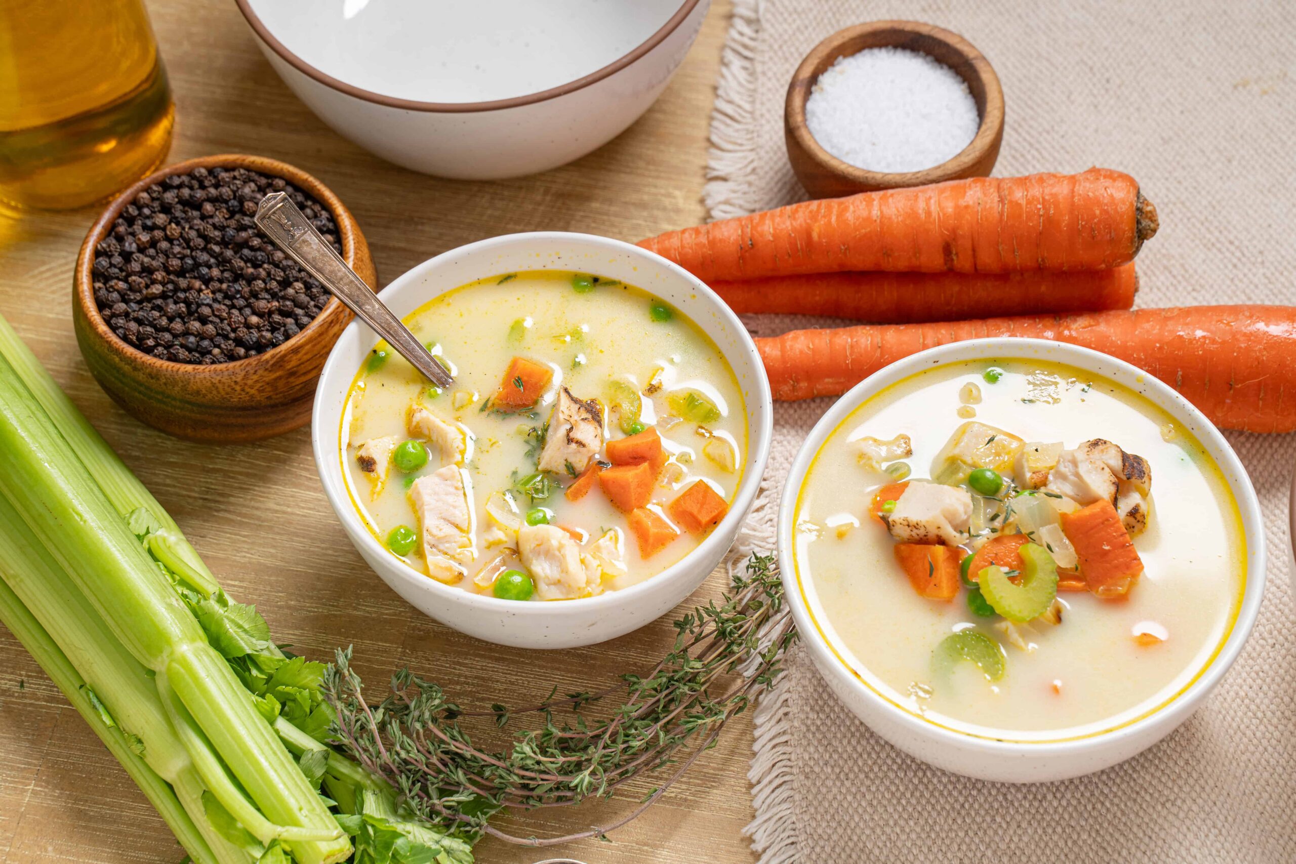 Savory chicken vegetable soup bowls with fresh carrots, celery, and herbs, seasoned with salt and pepper, featuring ingredients like olive oil and peas, served in white bowls on a wooden table.