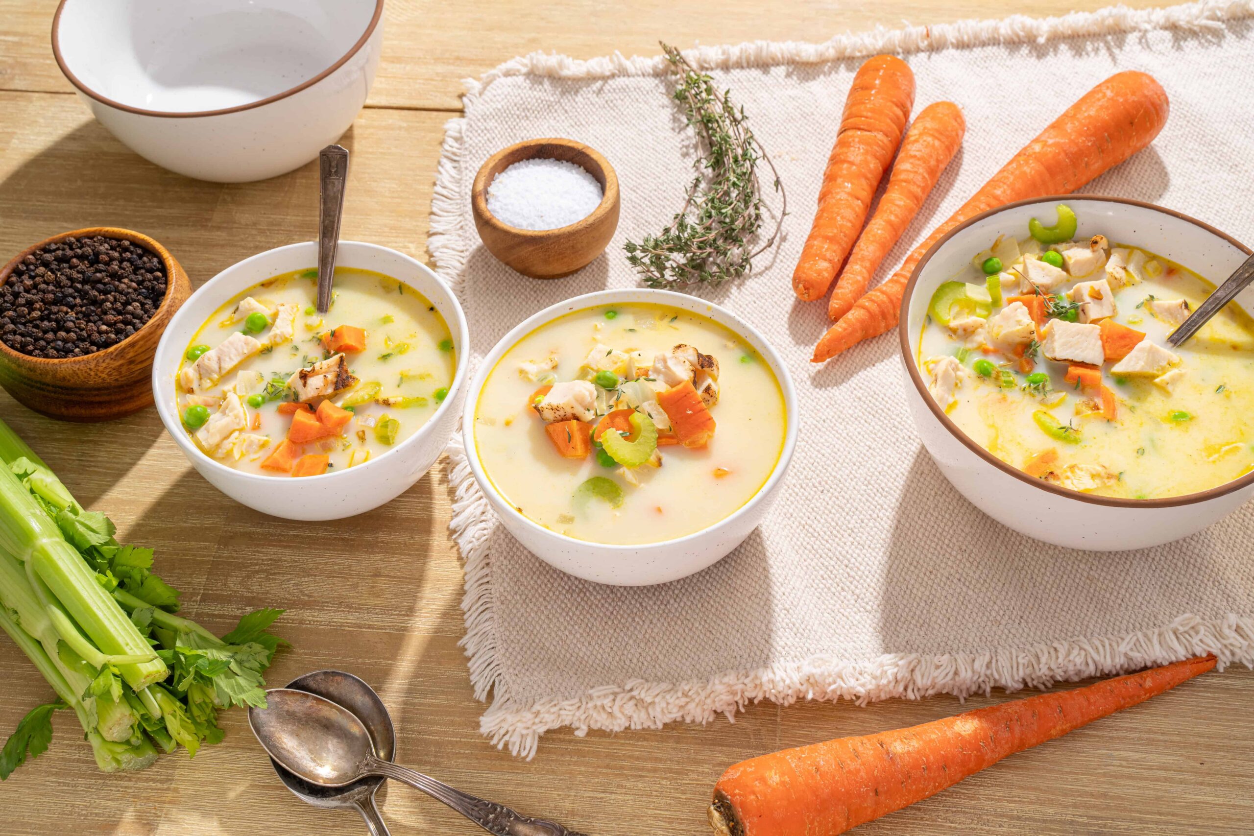 Creamy chicken and vegetable soup with carrots, celery, and peas in white bowls, surrounded by fresh ingredients like whole carrots, celery stalks, thyme, salt, and pepper on a wooden table.