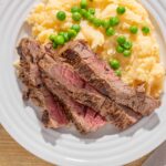 Juicy beef steak with mashed potatoes and green peas on a white plate, served with cutlery, perfect for a hearty dinner or lunch.