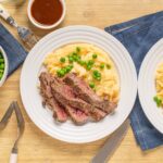 Juicy steak with mashed potatoes and green peas on a white plate, served with gravy in a small bowl, on wooden table with blue napkin, perfect comfort food for dinner.