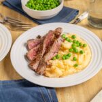 Juicy grilled steak with mashed potatoes and green peas on a white plate, served with gravy, on a wooden table for a delicious homemade dinner.