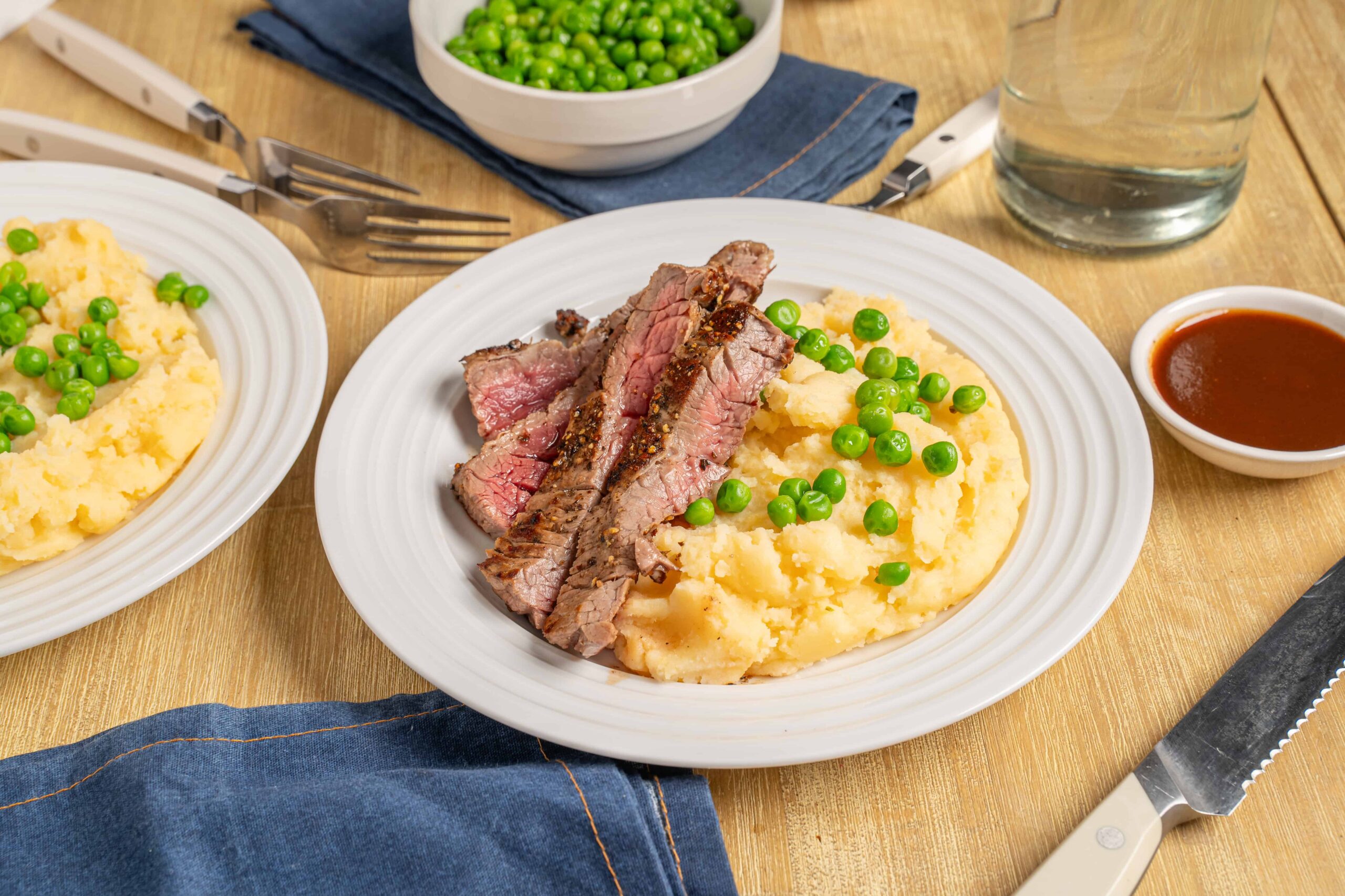 Juicy grilled steak with mashed potatoes and green peas on a white plate, served with gravy, on a wooden table for a delicious homemade dinner.