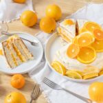 Juicy orange-flavored layered cake topped with fresh orange slices, served on a white plate with a slice on a separate plate, surrounded by whole oranges and white linen on a wooden table.