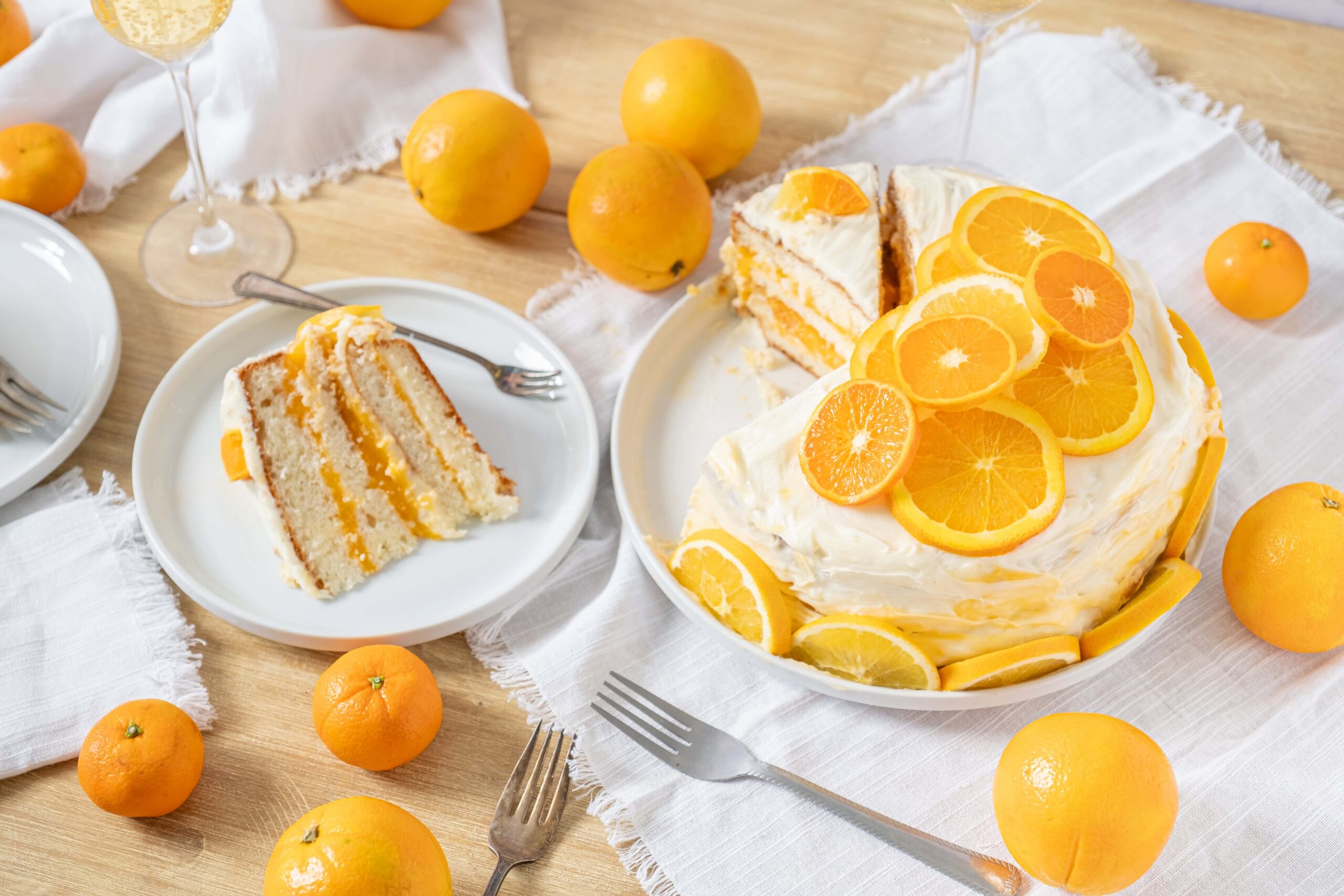 Juicy orange-flavored layered cake topped with fresh orange slices, served on a white plate with a slice on a separate plate, surrounded by whole oranges and white linen on a wooden table.