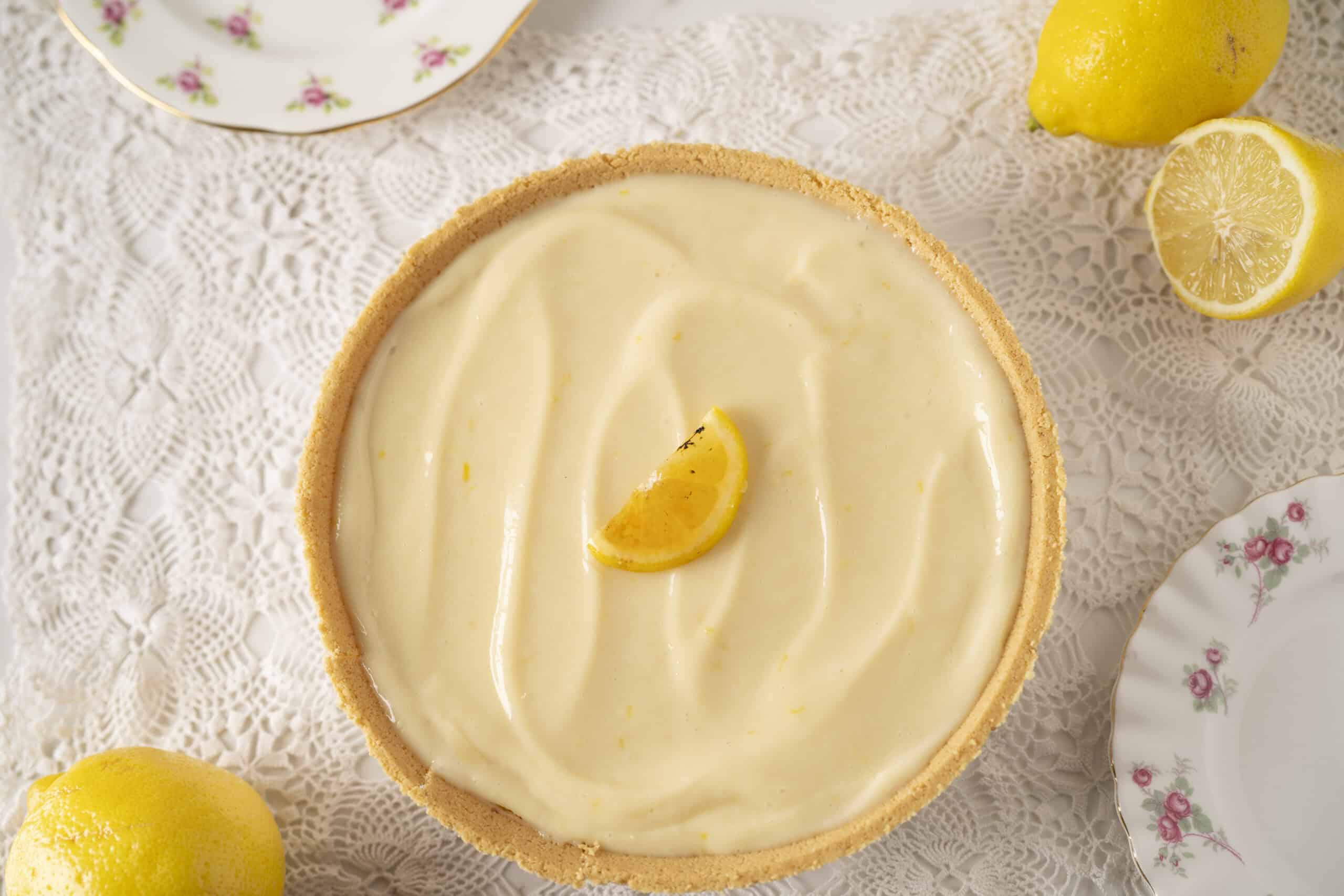 Lemon tart with a smooth, creamy lemon filling and a crumbly crust, garnished with a lemon wedge on top. Fresh lemons and vintage floral china add a charming touch to this classic dessert.