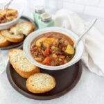 Savory vegetable chili served in a rustic bowl with slices of toasted bread on a dark plate, accompanied by a glass of water, salt and pepper shakers, and a soft white napkin, perfect for hearty comfort food.