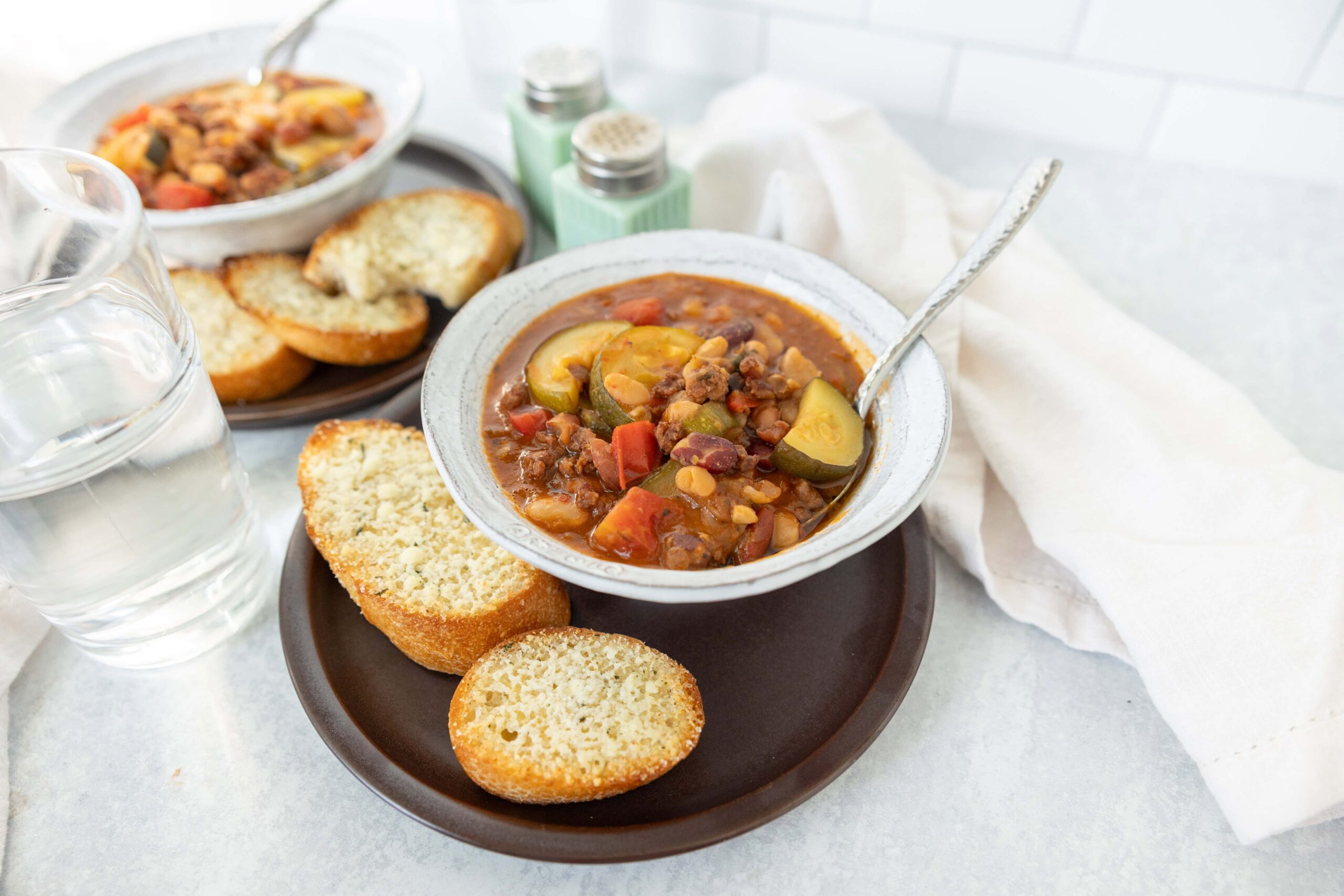 Savory vegetable chili served in a rustic bowl with slices of toasted bread on a dark plate, accompanied by a glass of water, salt and pepper shakers, and a soft white napkin, perfect for hearty comfort food.
