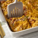 Creamy chicken casserole with shredded chicken, melted cheese, and mixed vegetables in a white baking dish, with a spatula ready to serve, on a light-colored table with gold-colored utensils.