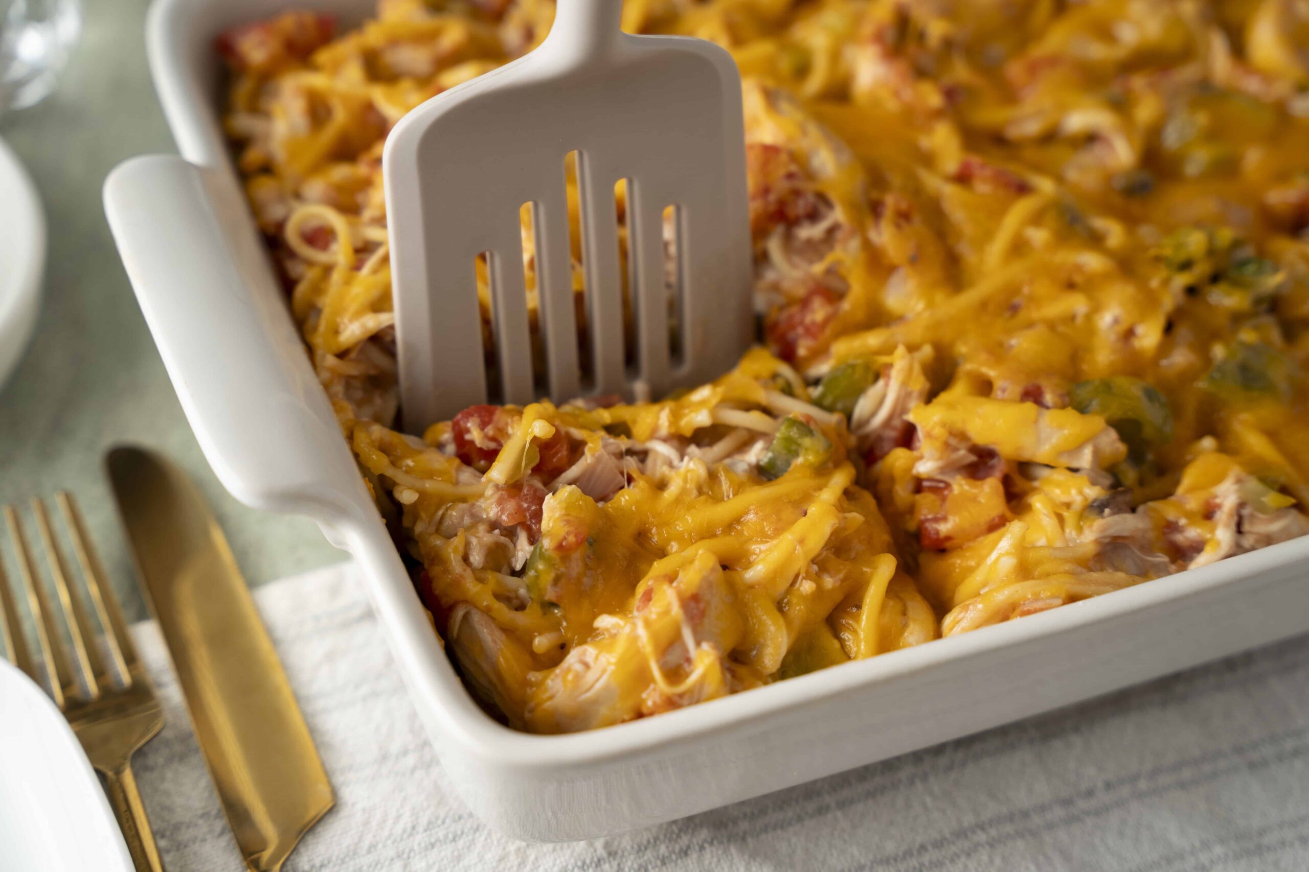 Creamy chicken casserole with shredded chicken, melted cheese, and mixed vegetables in a white baking dish, with a spatula ready to serve, on a light-colored table with gold-colored utensils.