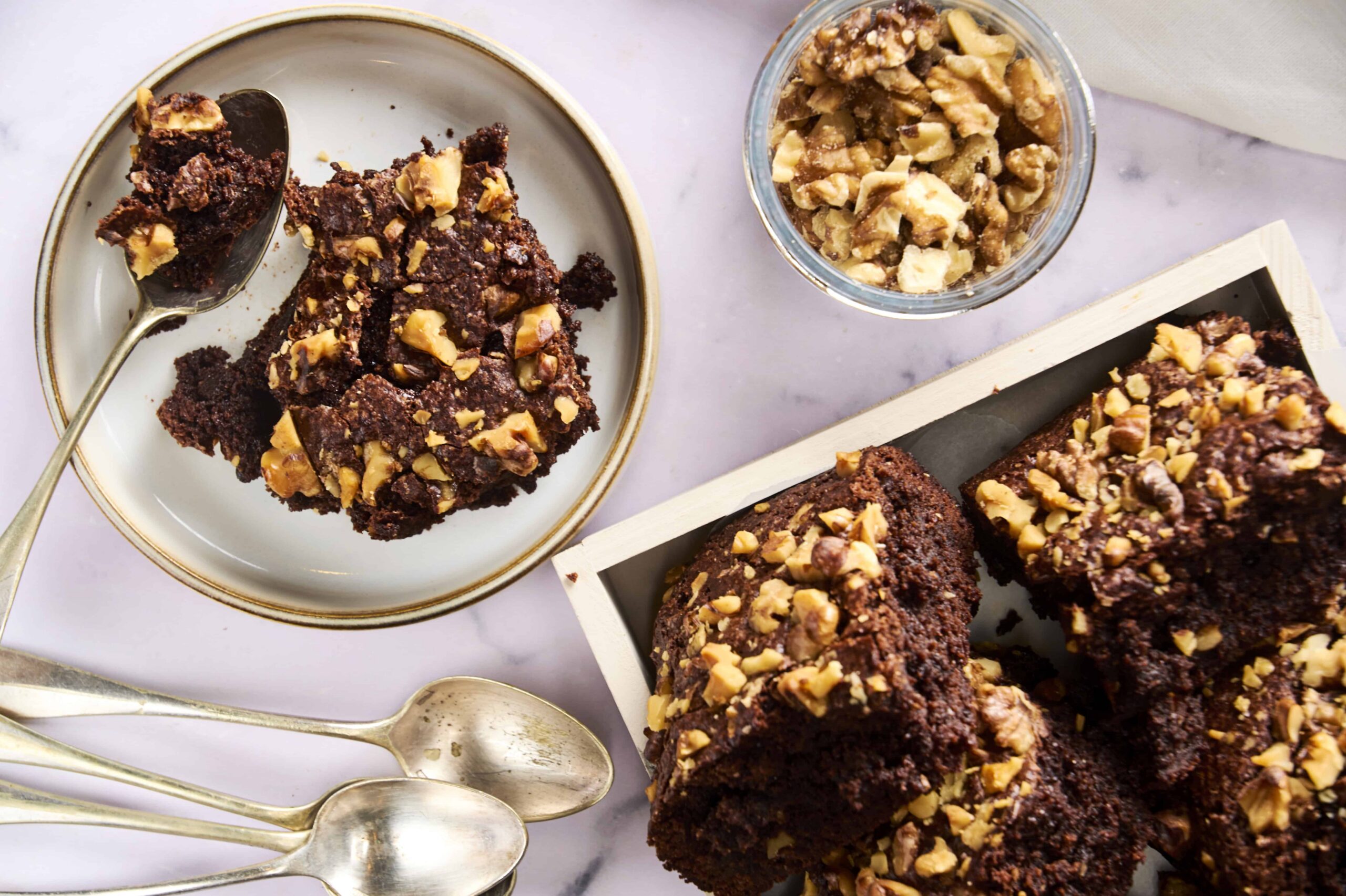 Rich chocolate walnut brownie with chopped walnuts on top, served on a white plate with a spoon, accompanied by more brownies and a jar of chopped walnuts, perfect for dessert or snack.