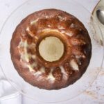 Golden-brown Bundt cake with a drizzle of glaze on a glass cake stand, served with vintage silver spoons on a decorative plate, perfect for dessert or special occasions.