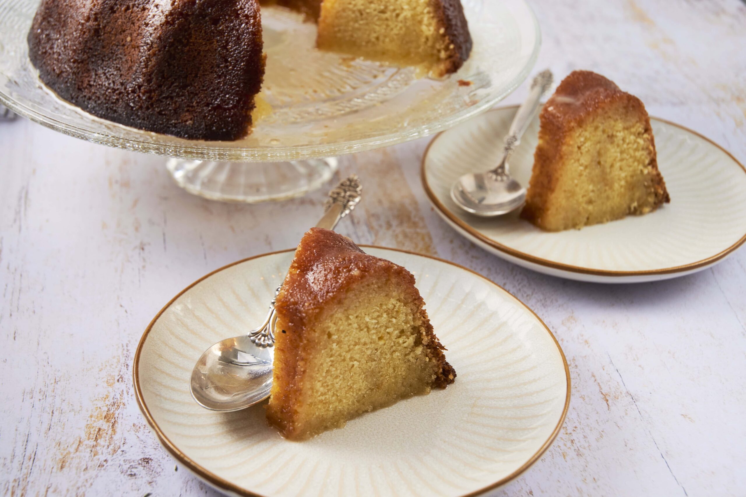 Moist vanilla pound cake slices on elegant white plates with gold rim, served with a glass cake stand of golden-brown crumb cake, perfect for dessert or tea time.