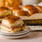 Golden brown breakfast sandwich with egg, ham, and cheese on a toasted bun with poppy seeds, served on a small plate in front of a tray of freshly baked bread rolls.