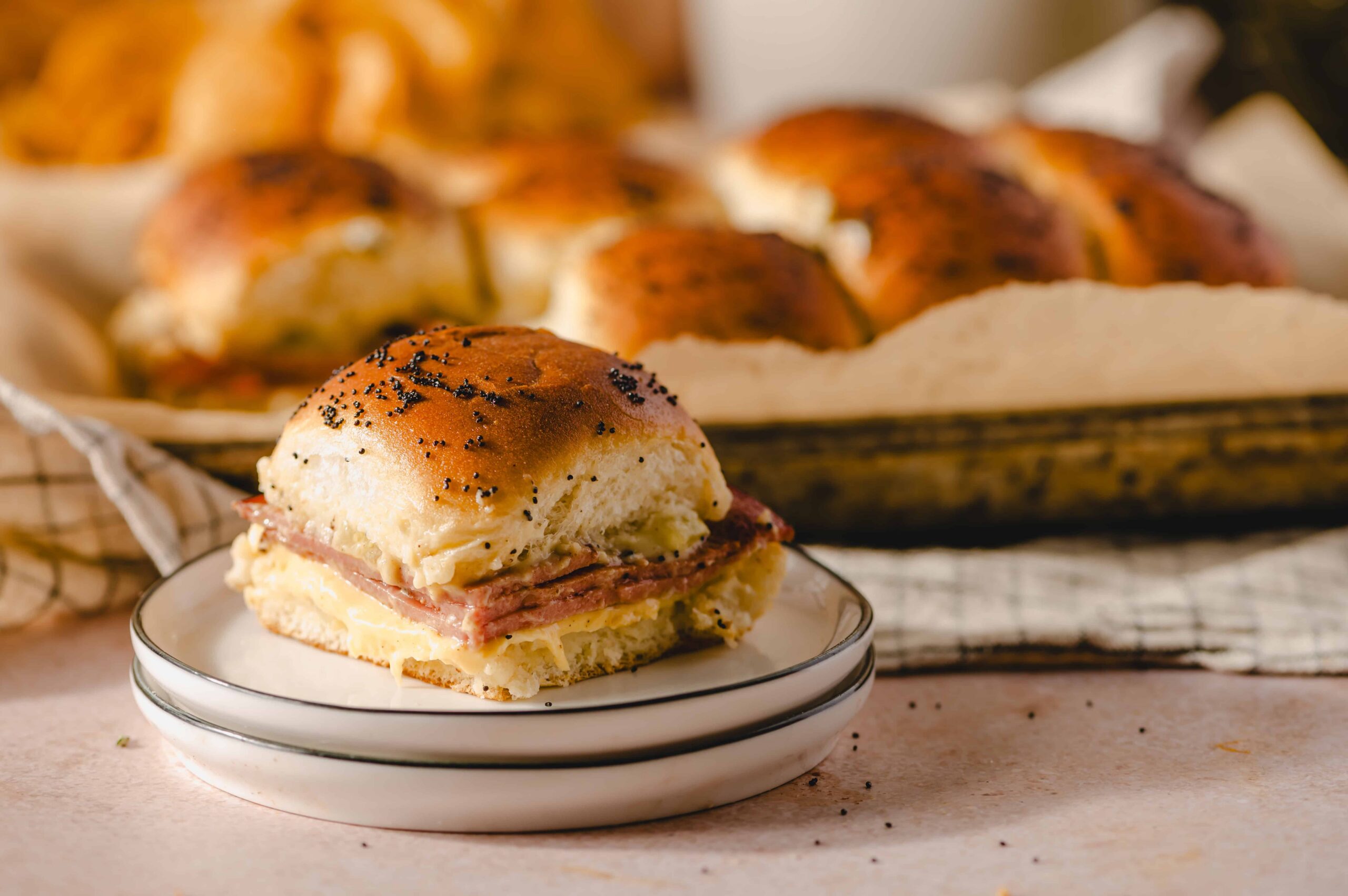 Golden brown breakfast sandwich with egg, ham, and cheese on a toasted bun with poppy seeds, served on a small plate in front of a tray of freshly baked bread rolls.