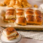 Golden brown ham and cheese sliders with poppy seeds on top, served on a baking sheet with crispy chips in the background, perfect for a delicious homemade appetizer or snack.