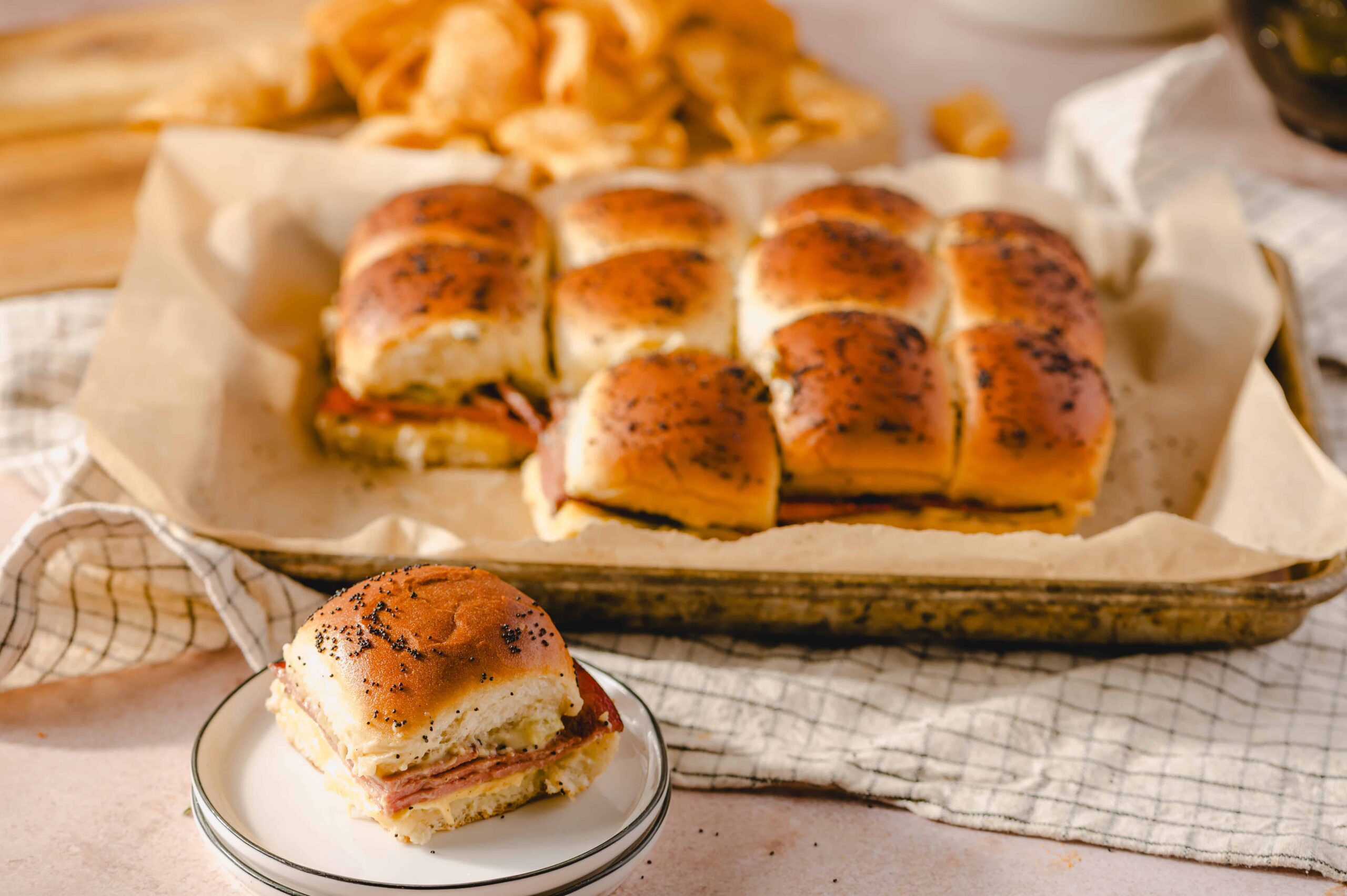 Golden brown ham and cheese sliders with poppy seeds on top, served on a baking sheet with crispy chips in the background, perfect for a delicious homemade appetizer or snack.