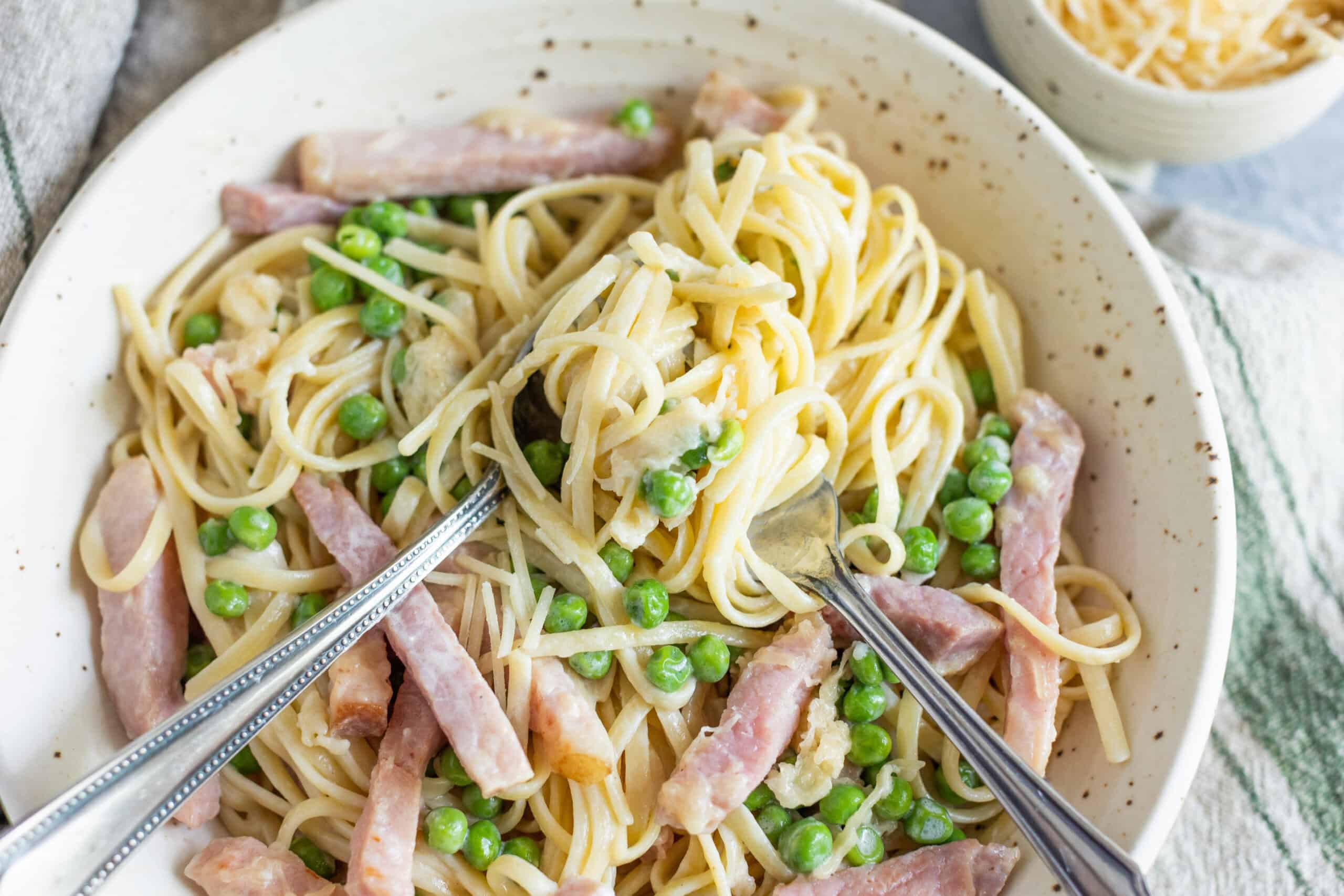 Creamy pasta with peas, ham, and cheese in a white bowl, topped with grated cheese and served with forks on a rustic table setting for a delicious homemade meal.