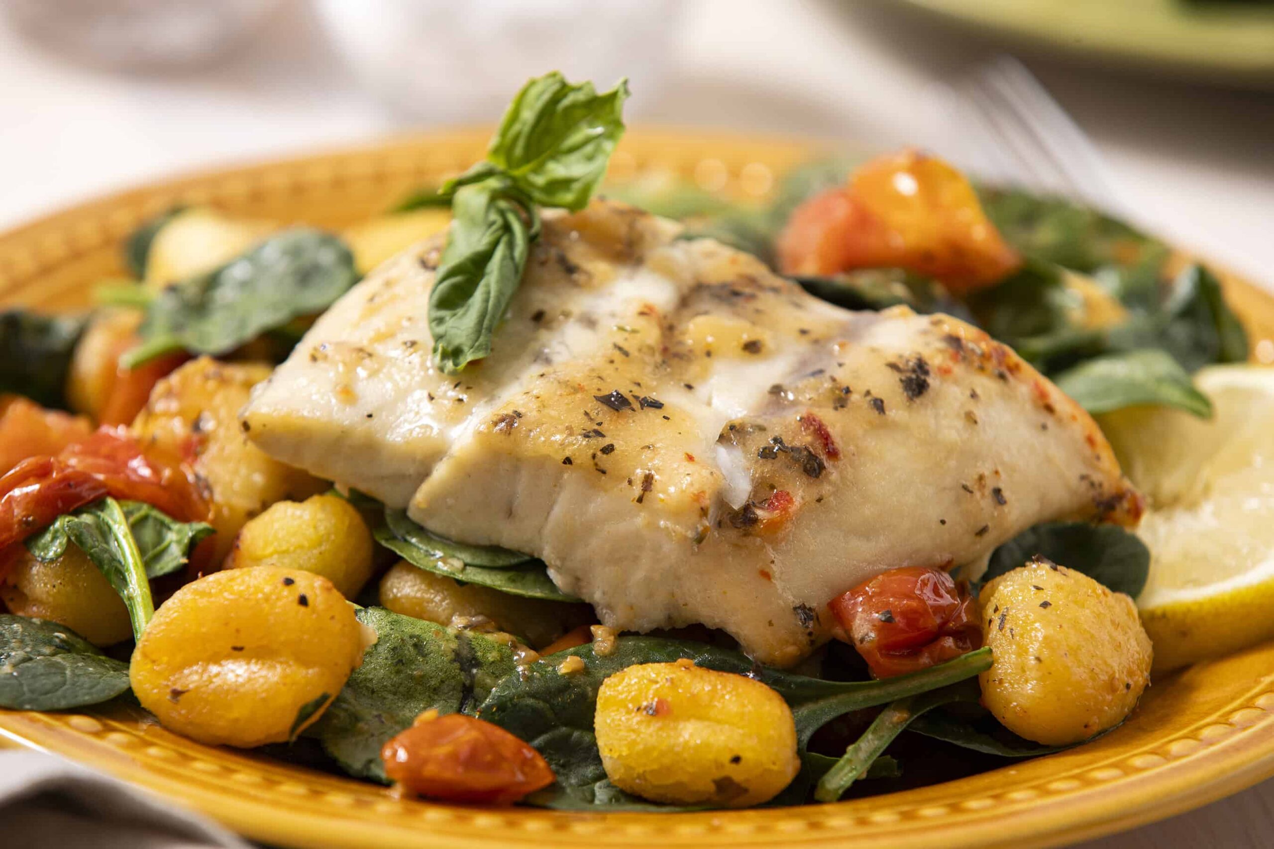 Fresh baked herb-crusted fish fillet with roasted potatoes, cherry tomatoes, spinach, and basil on a yellow ceramic plate. Perfect for a healthy, flavorful seafood dinner.