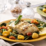 Fresh baked white fish fillet with lemon, baby potatoes, spinach, cherry tomatoes, and herbs on a yellow plate, served with lemon wedges, garnished with basil, perfect for healthy seafood recipes.