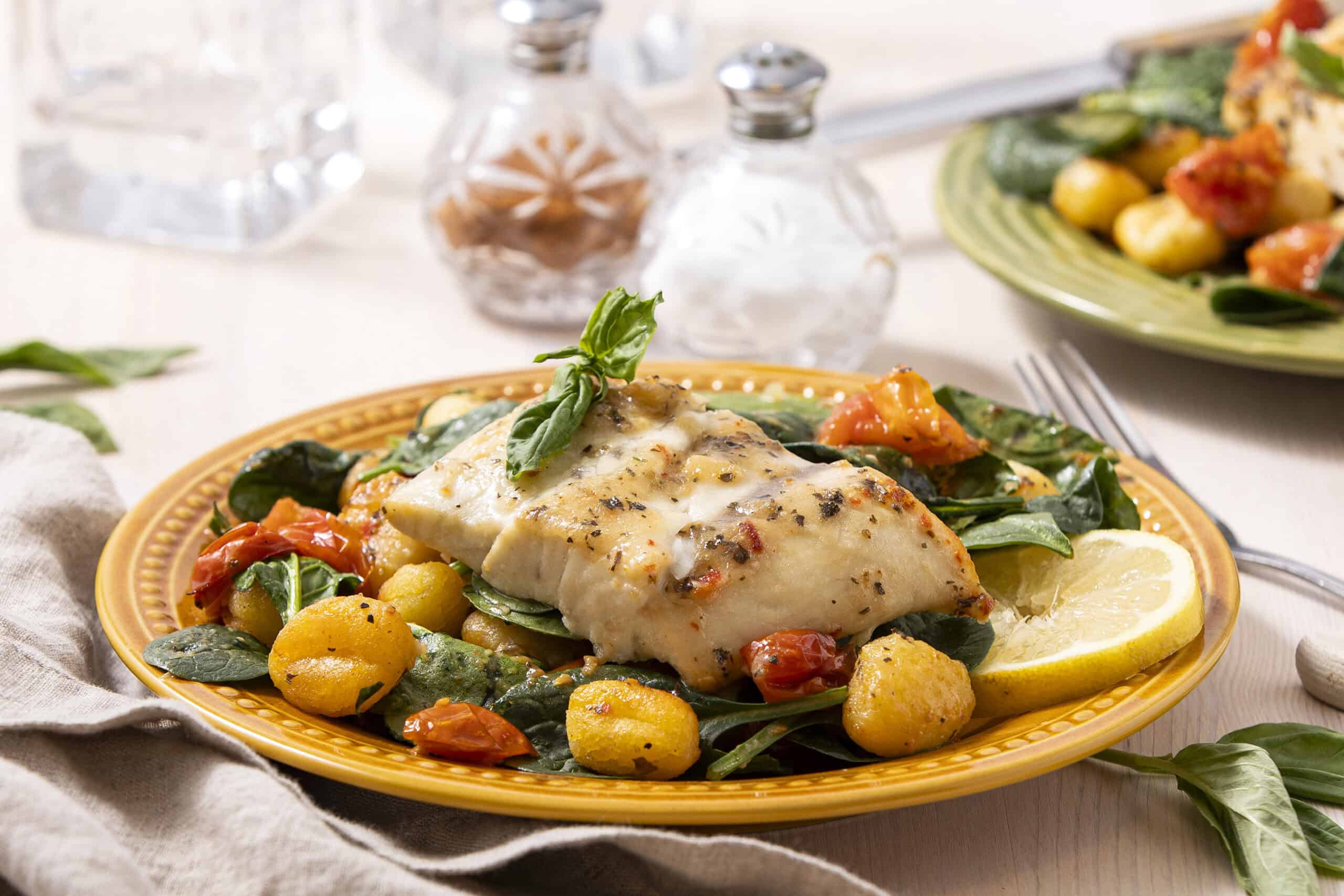 Fresh baked white fish fillet with lemon, baby potatoes, spinach, cherry tomatoes, and herbs on a yellow plate, served with lemon wedges, garnished with basil, perfect for healthy seafood recipes.