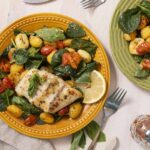 Fresh baked fish with lemon, served with spinach, cherry tomatoes, and gnocchi on colorful ceramic plates, garnished with herbs for a flavorful, healthy meal.