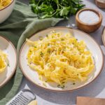 Creamy homemade fettuccine pasta with grated cheese, parsley, salt, and black pepper on white plates, served with fresh herbs and kitchen utensils for delicious Italian cuisine.