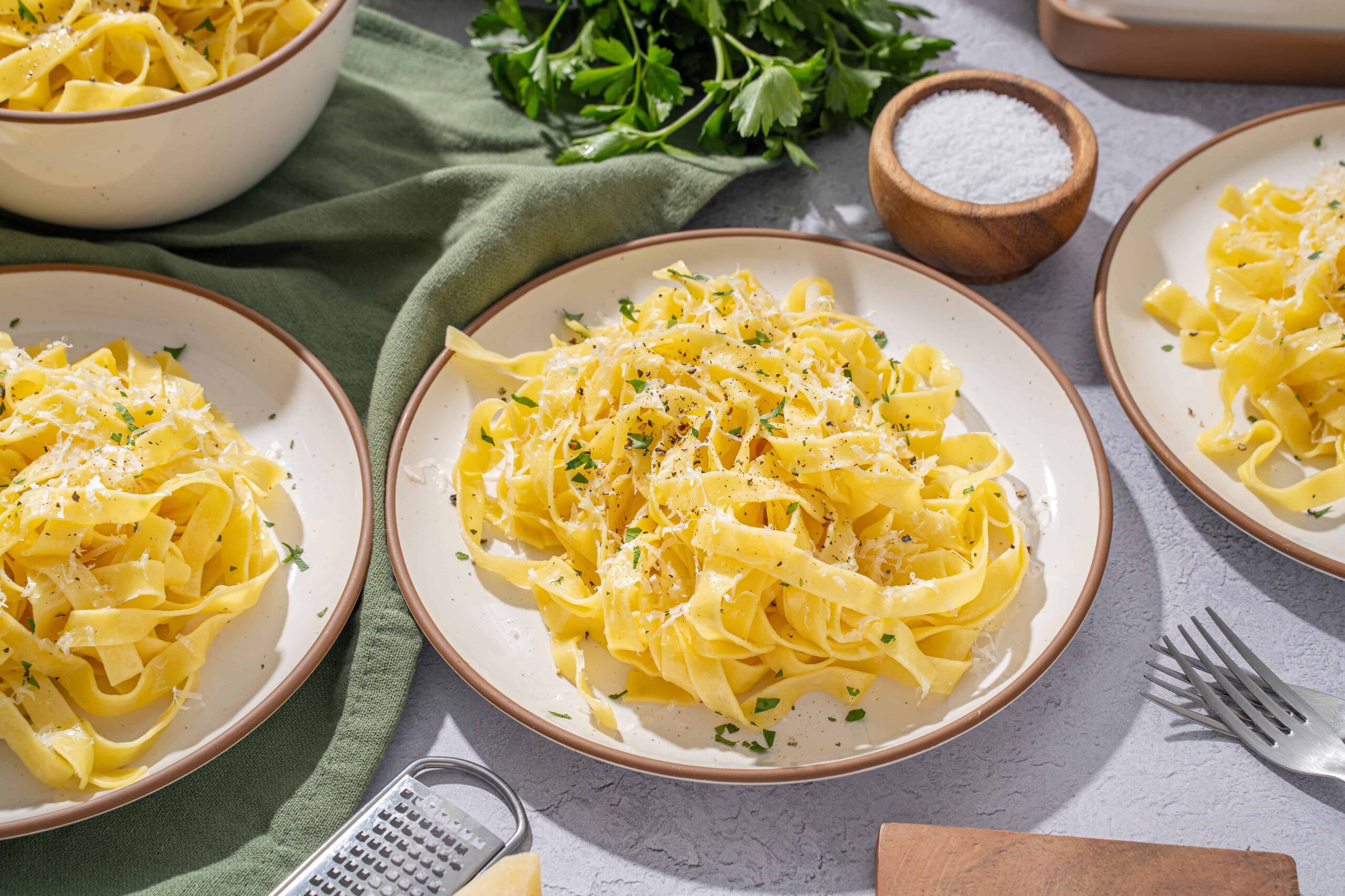 Creamy homemade fettuccine pasta with grated cheese, parsley, salt, and black pepper on white plates, served with fresh herbs and kitchen utensils for delicious Italian cuisine.