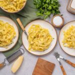 Creamy fettuccine alfredo pasta served on white plates with grated cheese and chopped parsley, garnished for a delicious Italian meal, on a textured gray surface with wooden accents and fresh herbs.