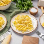 Creamy fettuccine alfredo pasta dish on white plates with grated Parmesan cheese, served with fresh parsley, salt, and black pepper on a light gray textured surface, perfect for Italian comfort food.