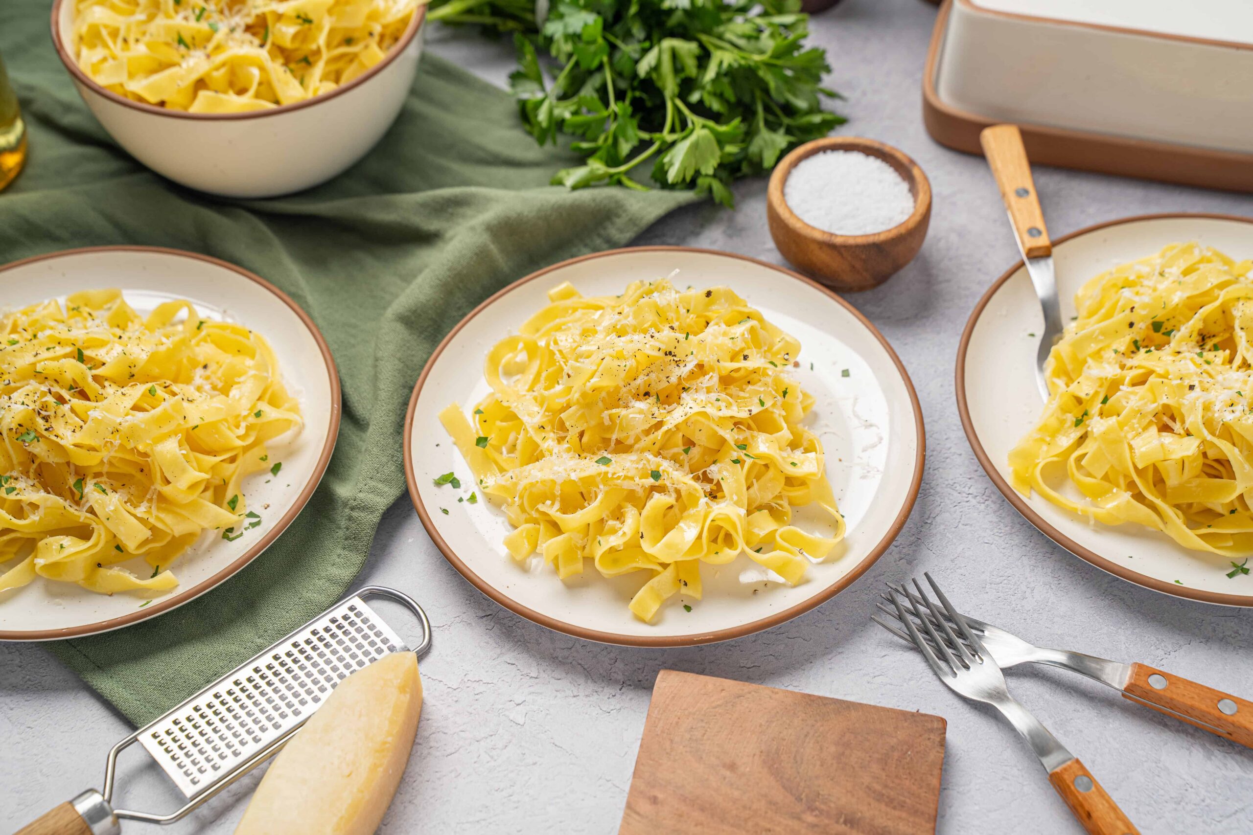 Creamy fettuccine alfredo pasta dish on white plates with grated Parmesan cheese, served with fresh parsley, salt, and black pepper on a light gray textured surface, perfect for Italian comfort food.