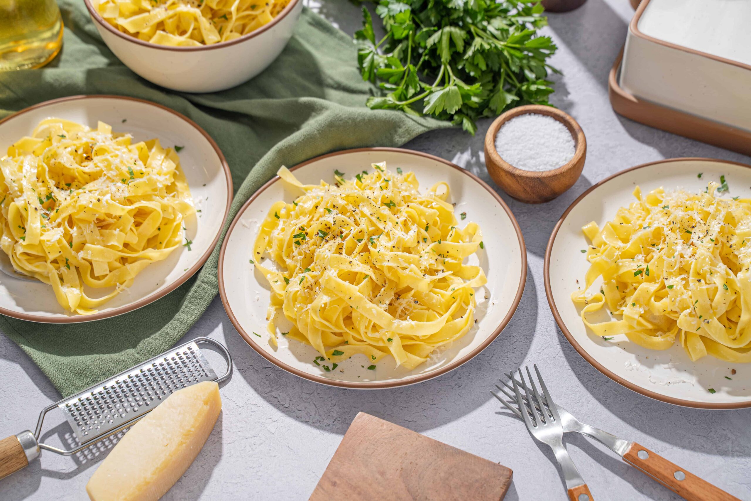 Creamy homemade fettuccine Alfredo with fresh parsley, served on rustic plates, with ingredients like Parmesan cheese, salt, and pasta on a kitchen countertop.