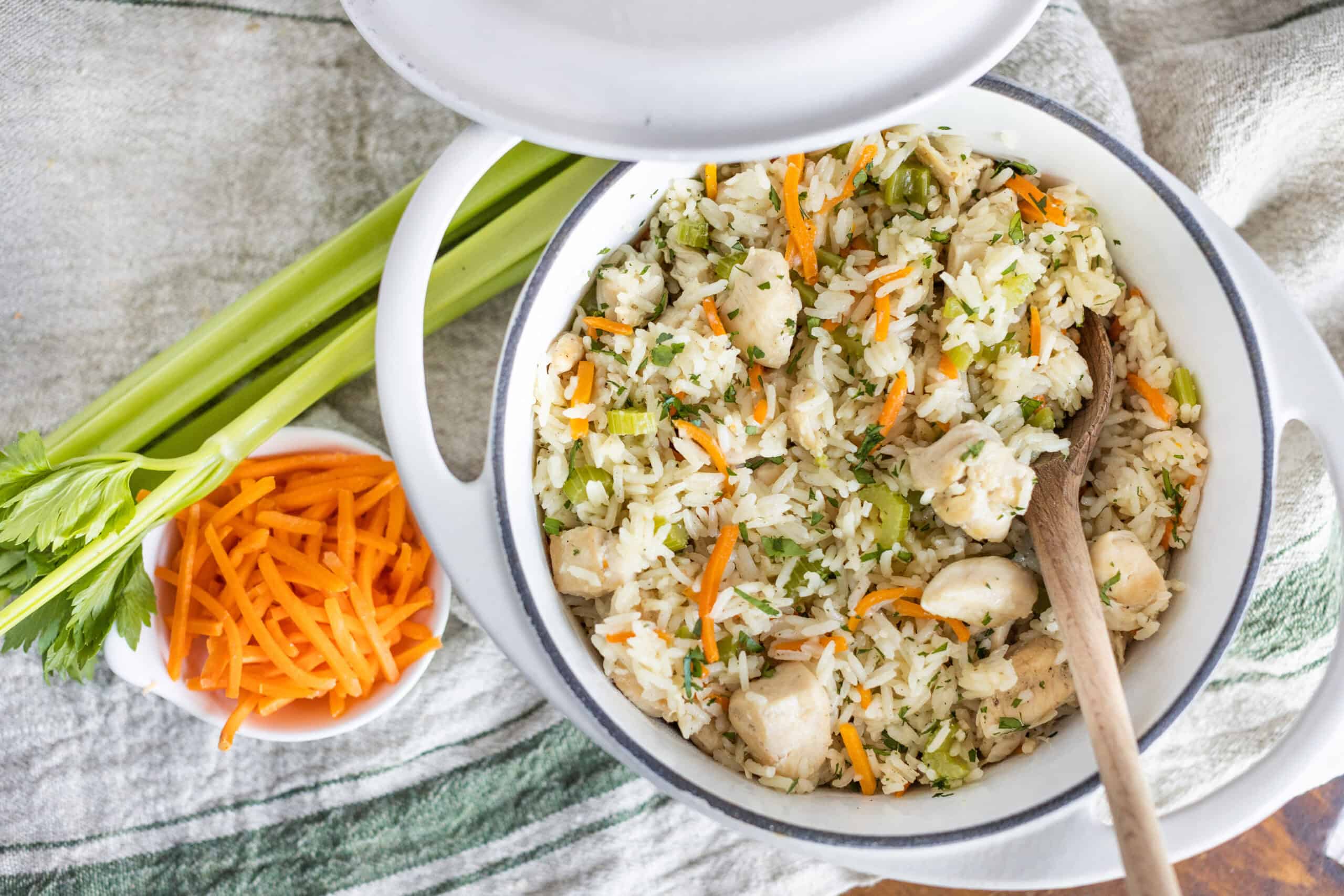 Creamy chicken fried rice with vegetables served in a white bowl, garnished with fresh herbs, surrounded by celery stalks, shredded carrots, and fresh cilantro for a flavorful Asian-inspired dish.