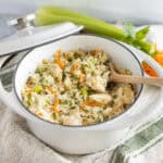 Steamed chicken rice in a white enamel pot with vegetables and herbs, on a rustic cloth with fresh celery and shredded carrots in the background.